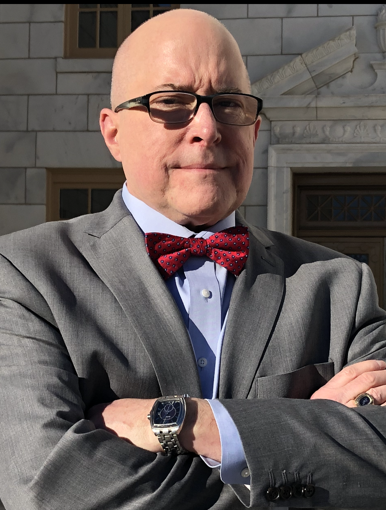 Man in gray suit with red bow tie and glasses crossing arms.