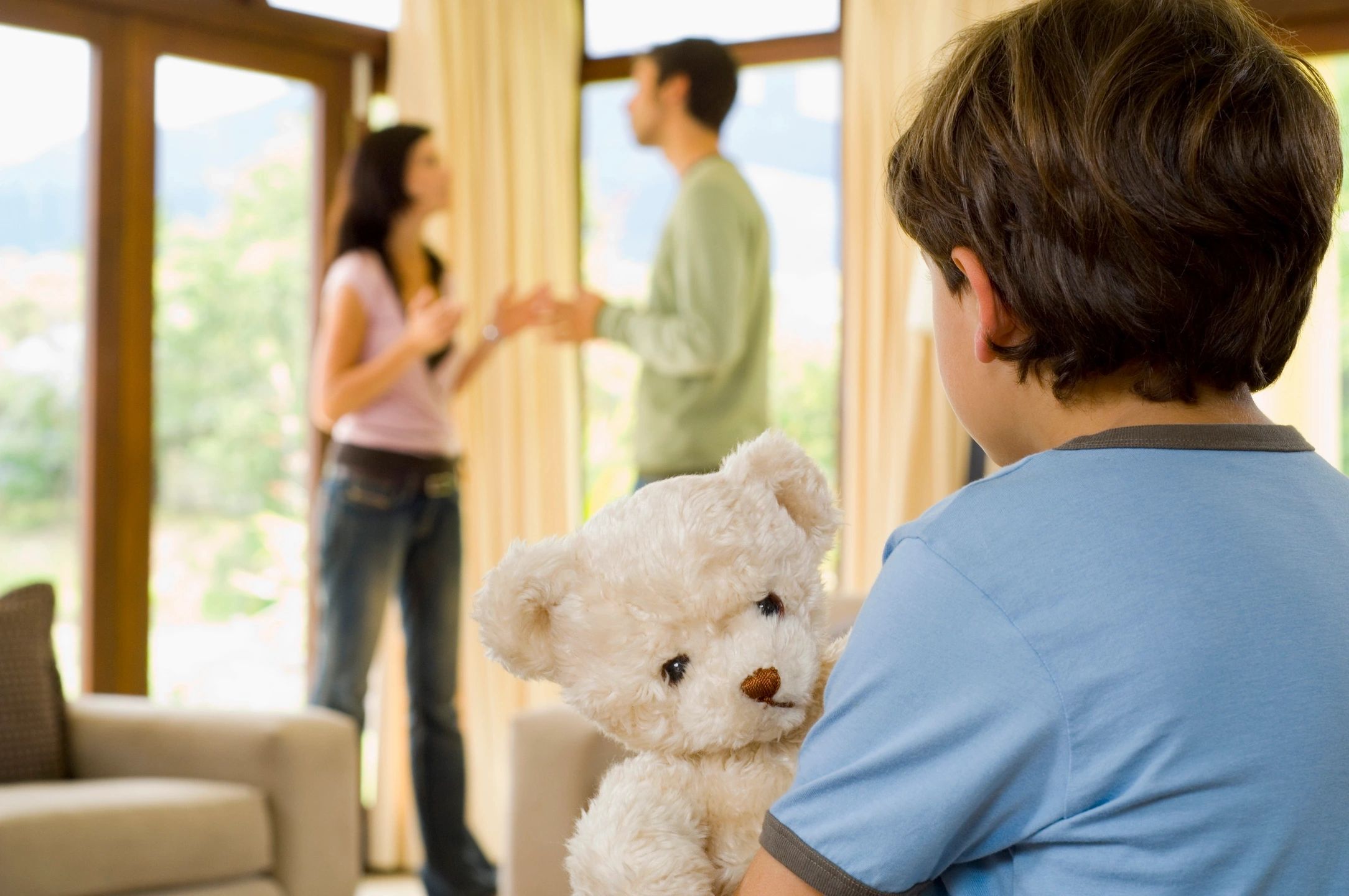 Child holding a teddy bear, watching parents argue.
