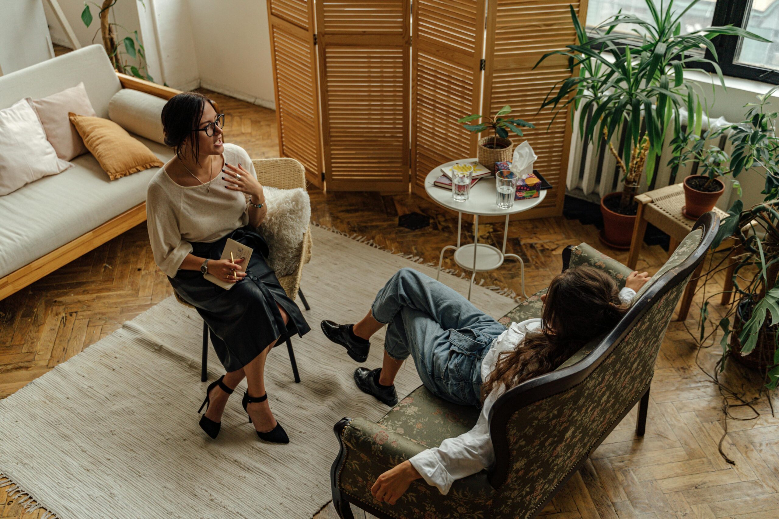 Two women engaged in a relaxed conversation in a cozy, plant-filled room.
