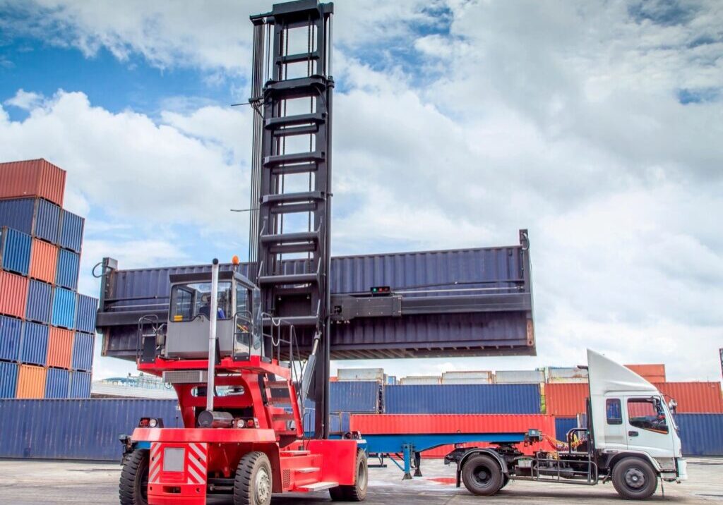 A large red container handler lifting a shipping container outdoors.