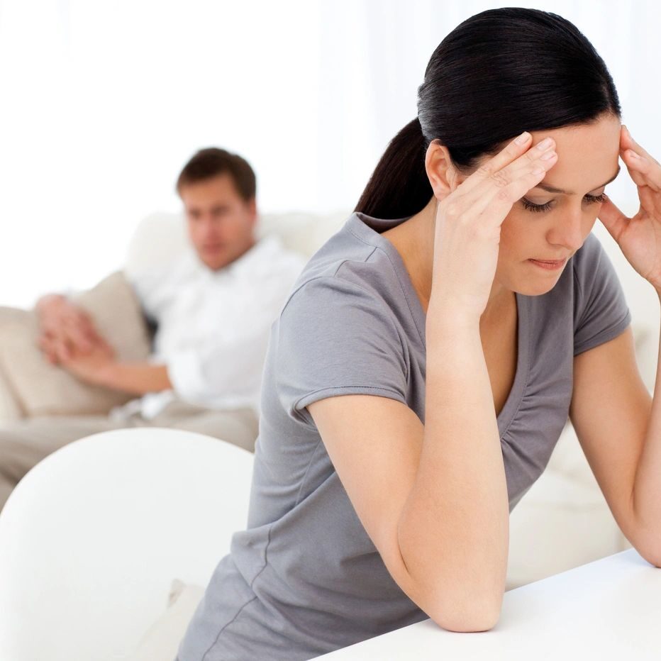 Woman stressed, man sitting in background, possibly tense relationship.
