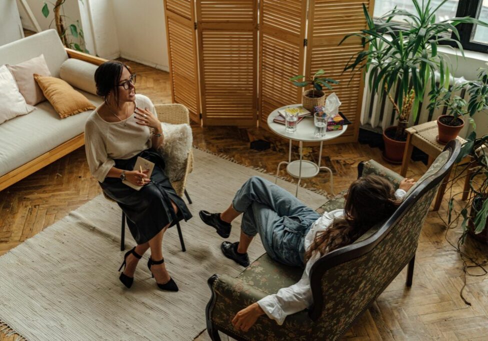 Two women engaged in a relaxed conversation in a cozy, plant-filled room.