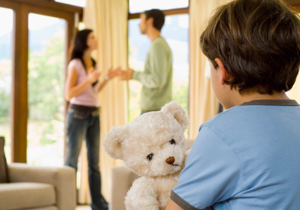 Child holding a teddy bear, watching parents argue.